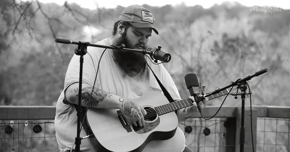 A man with a beard and tattoos plays an acoustic guitar while singing into a microphone.