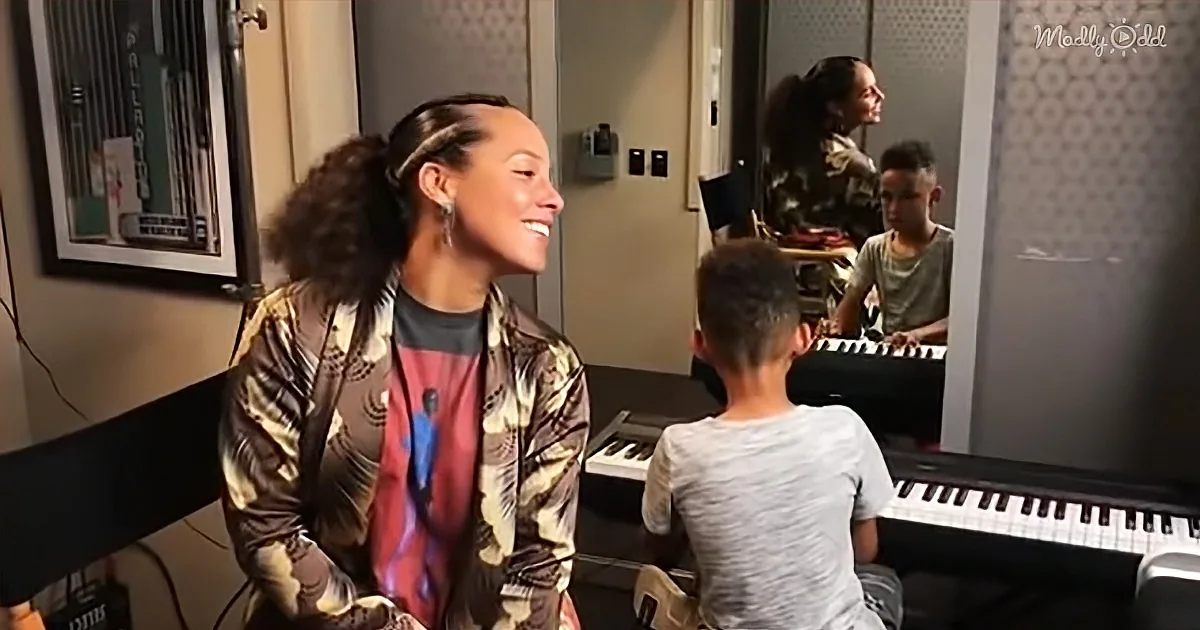 A woman in a camouflage jacket smiles as a young boy plays a keyboard in a music studio.