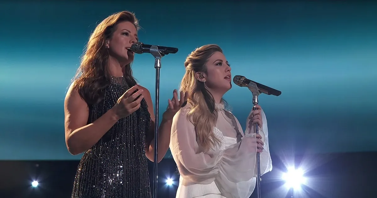 Two women sing on stage, one in black, the other in white, under bright lights.