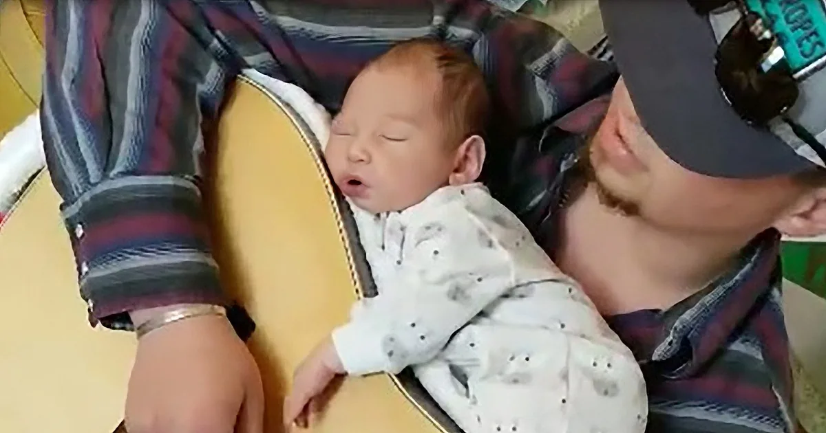 A baby in a white onesie sleeps peacefully in a yellow chair, held by a person in a plaid shirt and cap.