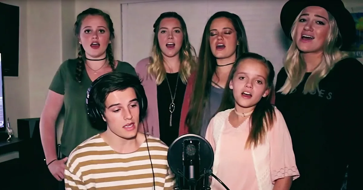 A group of six people, including a young man in a striped shirt, are singing in front of a microphone.