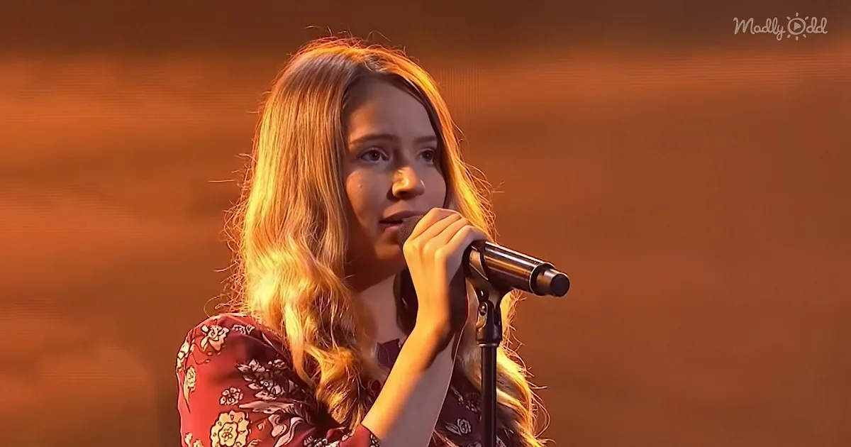 A person with long blonde hair sings into a microphone, wearing a floral top, against a warm, blurred background.