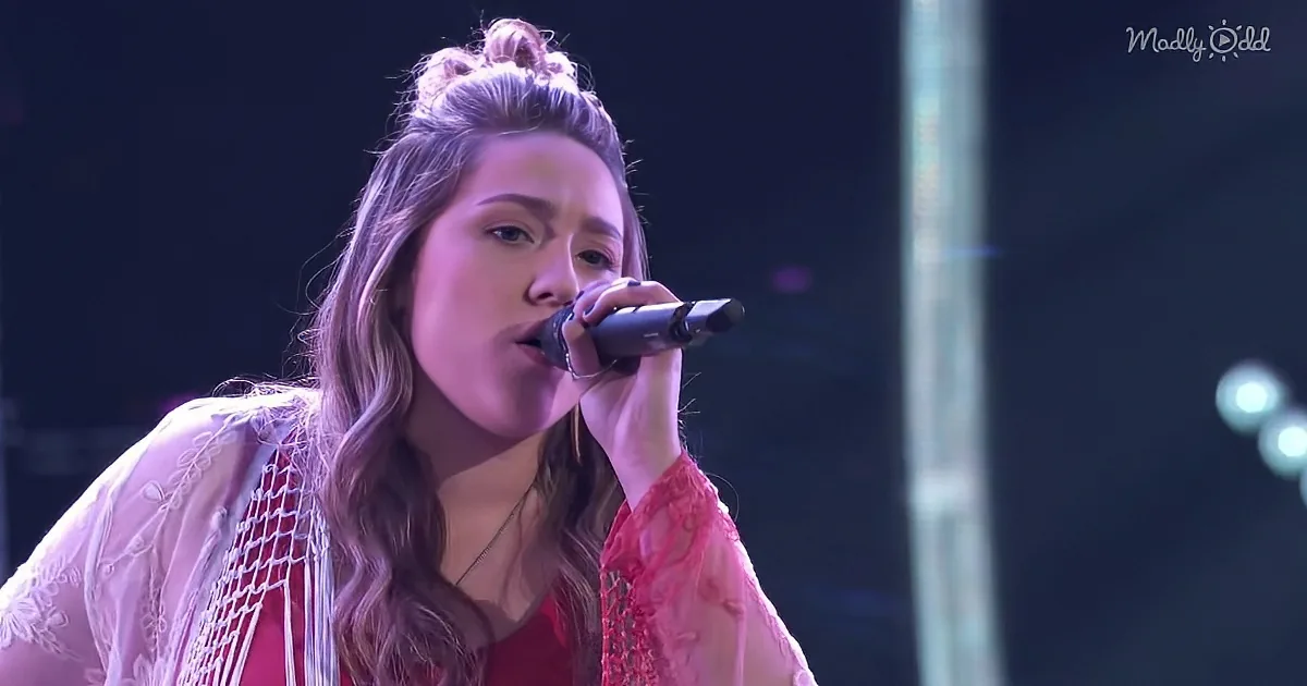 A woman with long hair, wearing a red top and white lace, sings into a microphone on stage.