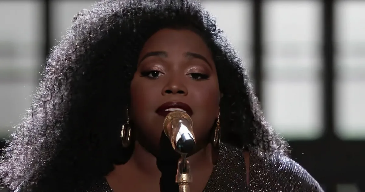 A woman with large, curly hair sings into a microphone, wearing a sparkly top and gold earrings.