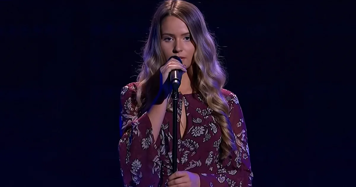 A woman with long, wavy hair in a floral dress sings into a microphone on stage.
