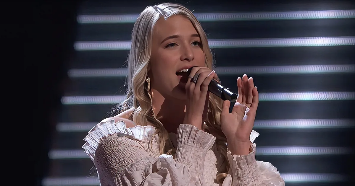 A woman with blonde hair in a white ruffled top sings into a microphone, hands raised, against a blurred light background.