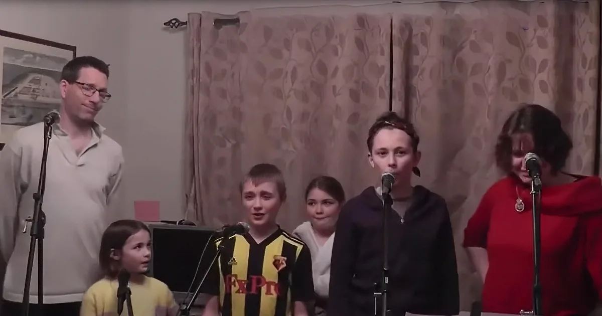 A group of children and an adult sing into microphones in a room with curtains.