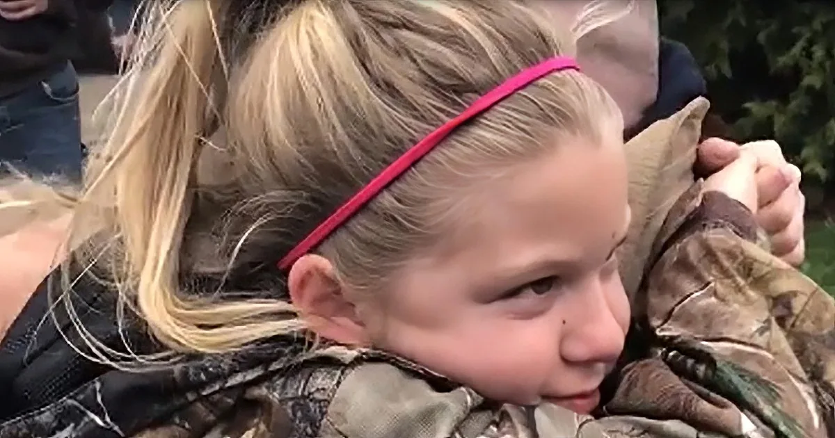 A young girl with blonde hair and a pink headband is being held by someone in camouflage clothing.