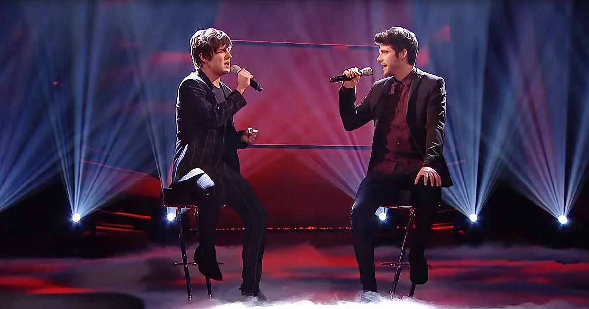 Two men in suits sing on stage with microphones, illuminated by red and blue lights.