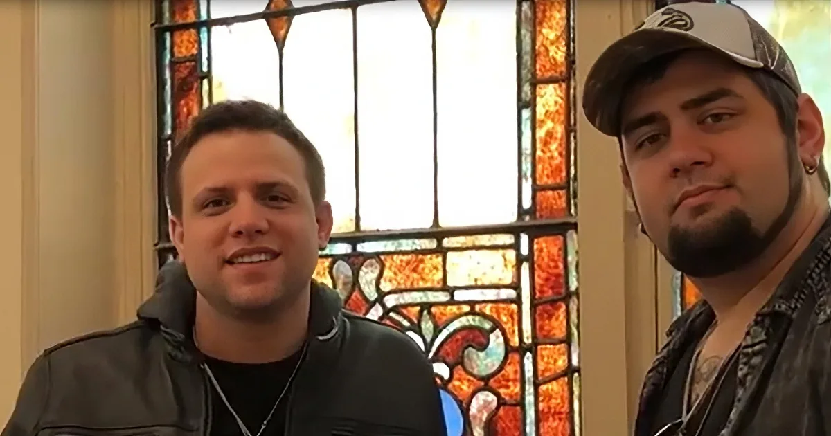 Two men, one smiling, one serious, in front of a stained glass window.