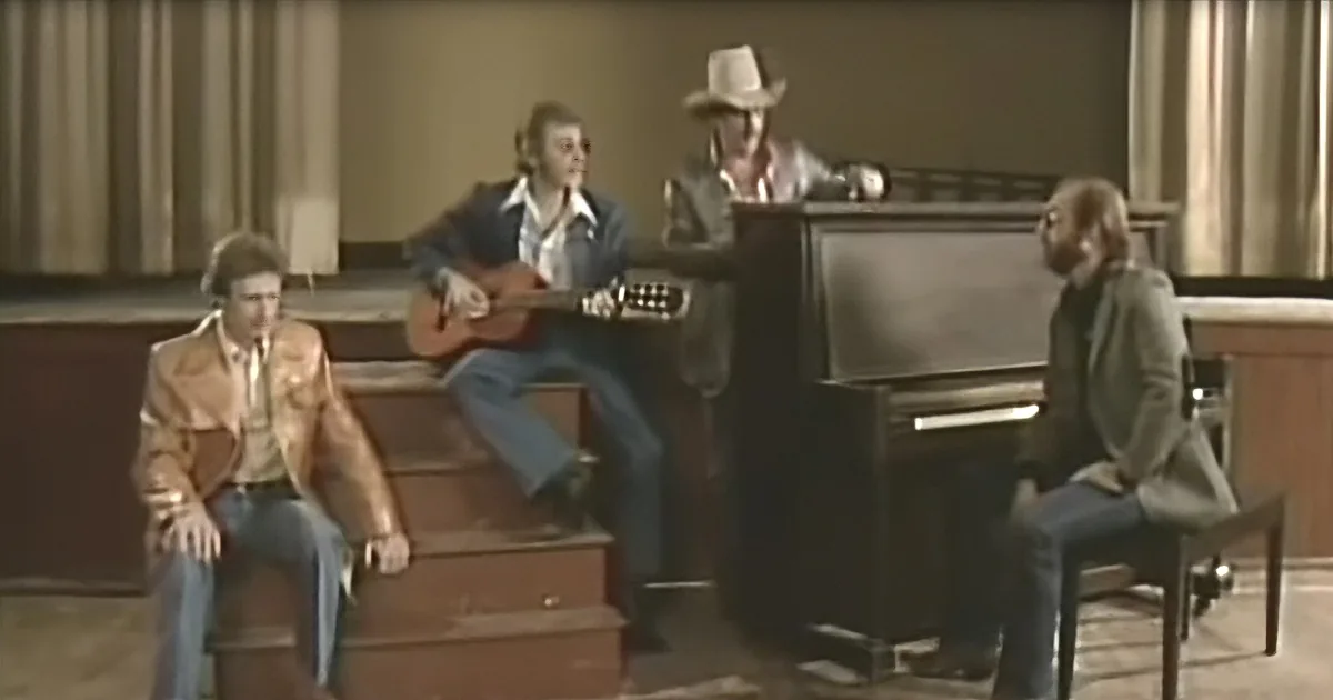 A man in a cowboy hat plays piano while another plays guitar, with two others seated nearby.