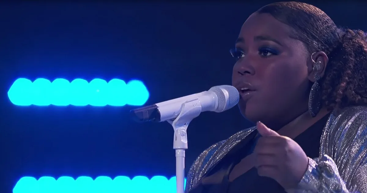 A woman sings into a microphone, giving a thumbs-up, with blue lights in the background.