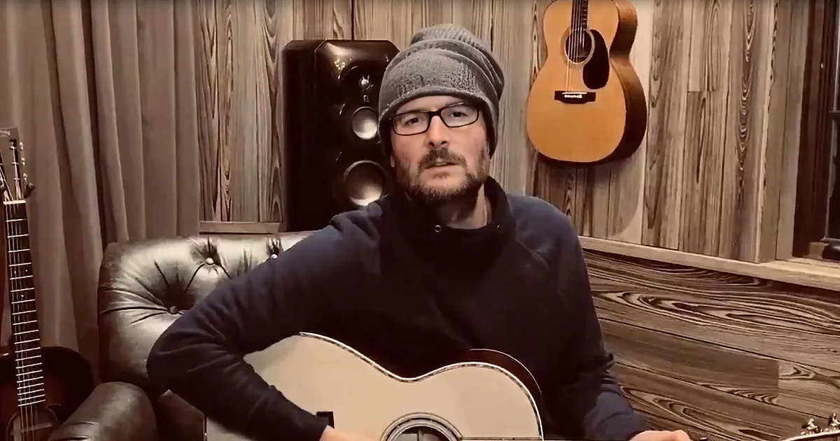 A man in a beanie and glasses sits on a leather couch, playing an acoustic guitar in a room with wood paneling and...