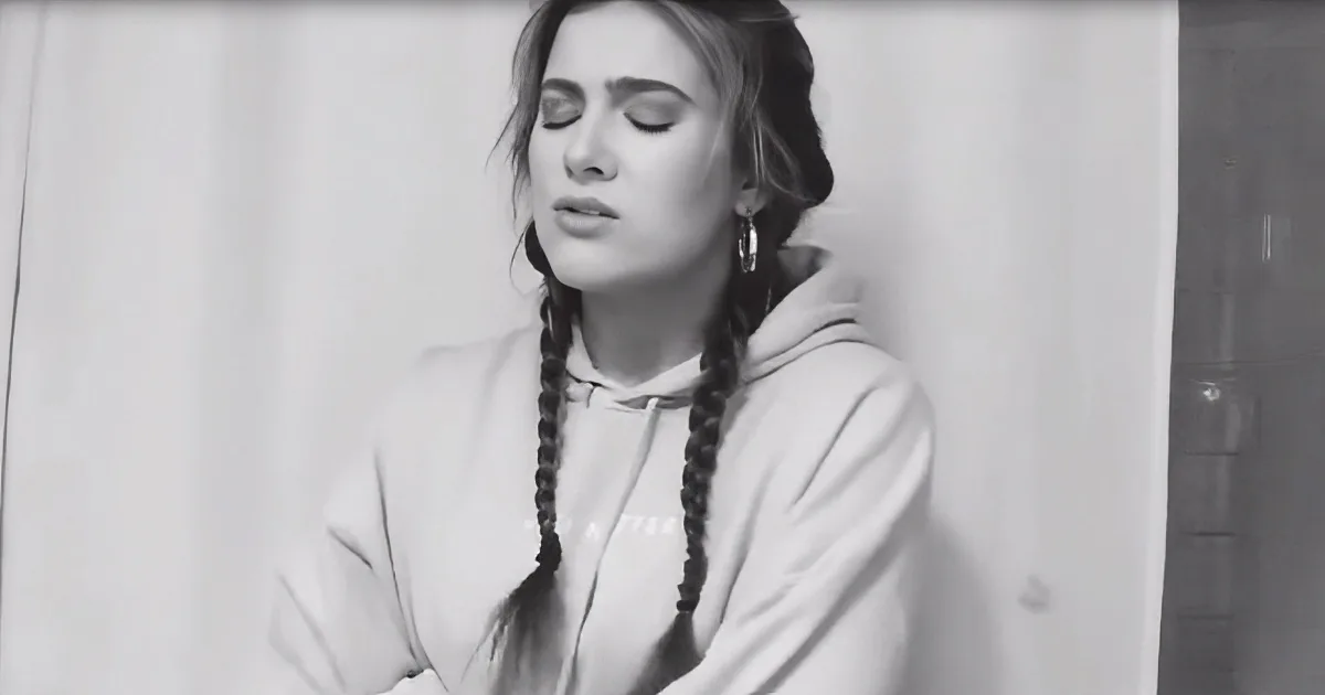 A woman with braided hair and hoop earrings, wearing a hoodie, stands with her eyes closed, appearing contemplative.