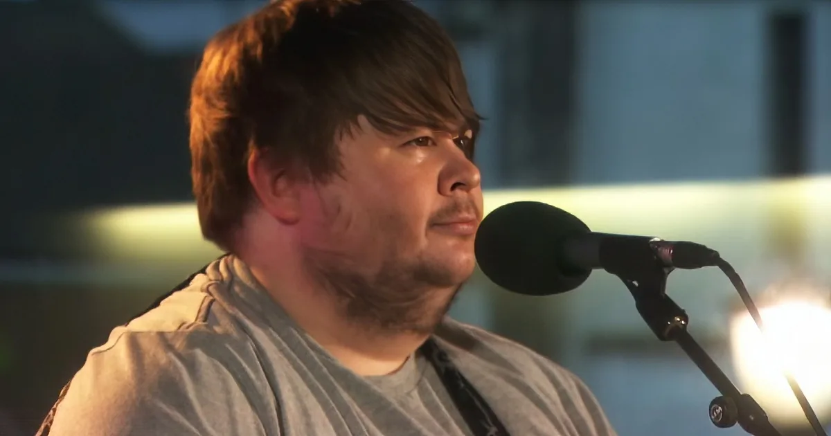A man with short brown hair and a beard is singing into a microphone.
