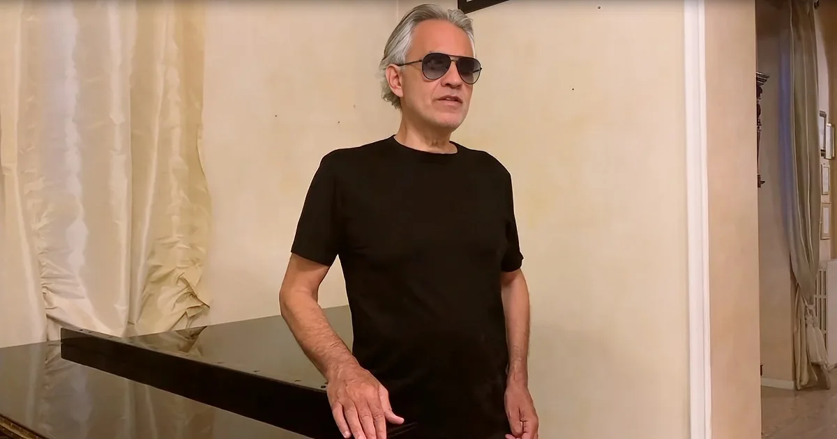 A man in a black shirt and sunglasses stands near a piano, looking to the side.
