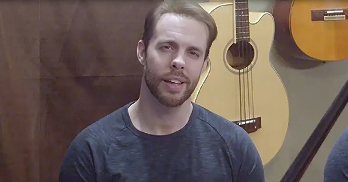 A man with a beard and short hair, wearing a dark t-shirt, stands in front of a guitar and a wooden wall.