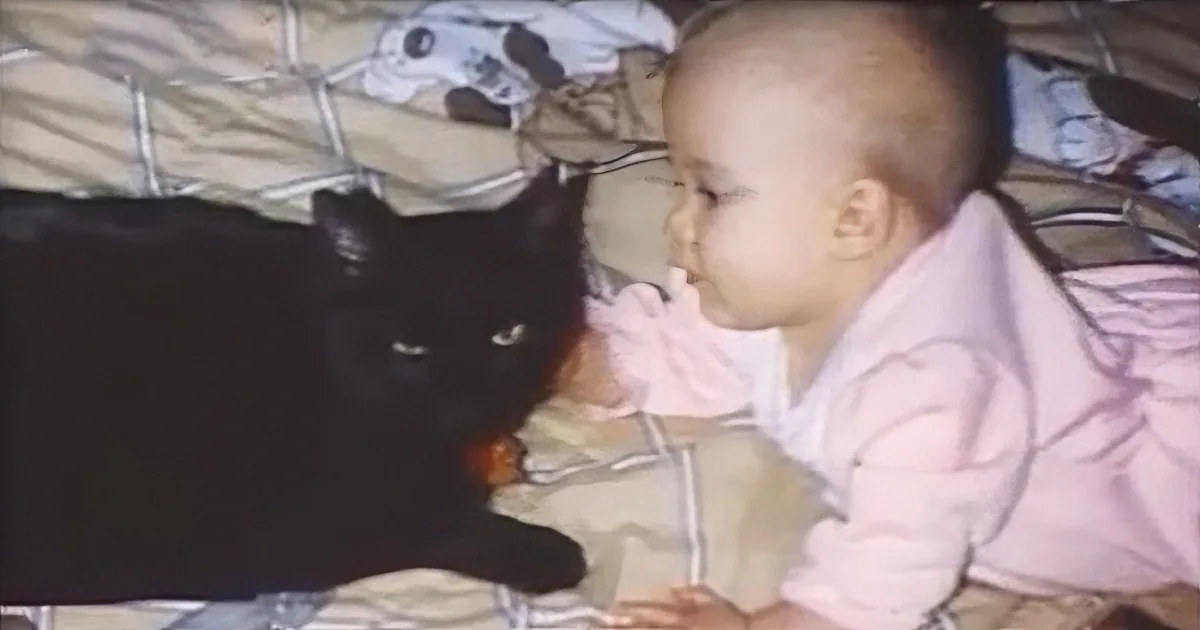 A baby in pink pajamas is lying on a bed, interacting with a black cat.
