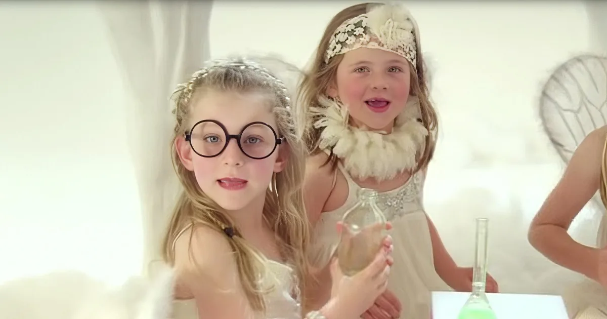 Two young girls in white dresses, one holding a bottle, another wearing a feather headband, in a bright, whimsical setting.