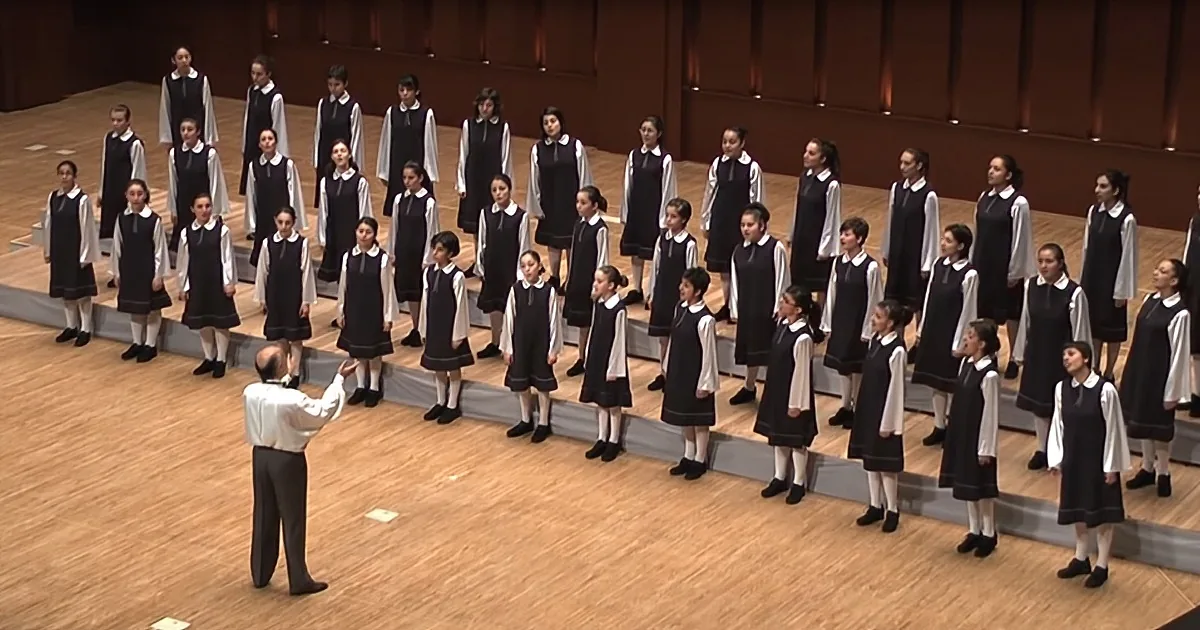 A conductor leads a choir of young girls in uniform on a stage.