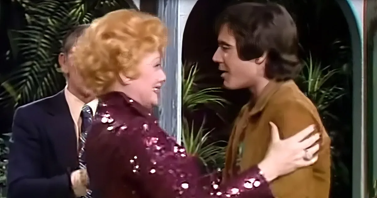 A woman in a sequined purple dress embraces a man in a brown jacket, both smiling, with greenery in the background.