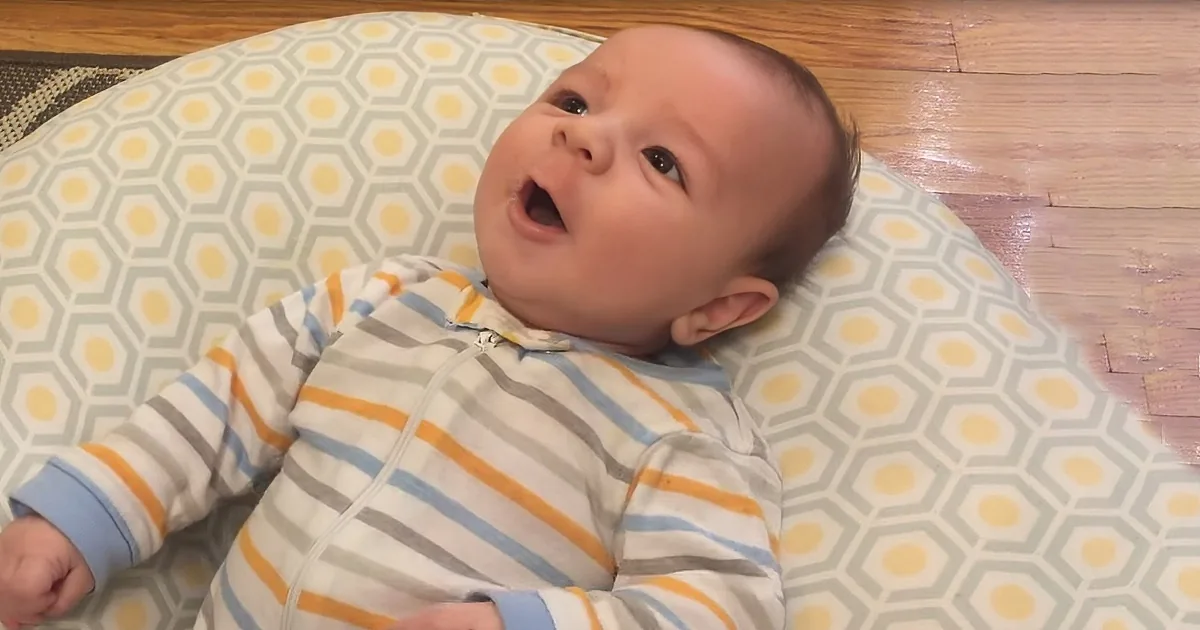 A baby in striped pajamas lies on a hexagonal-patterned cushion on a wooden floor.