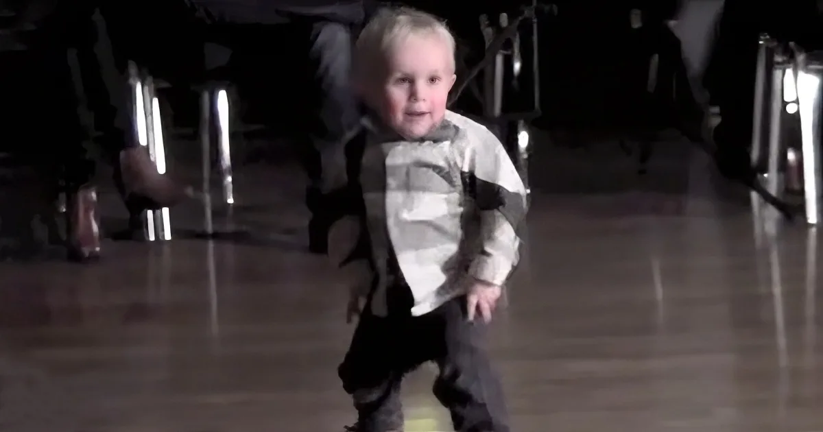 A young child in a striped shirt and dark pants is walking on a wooden floor, smiling.