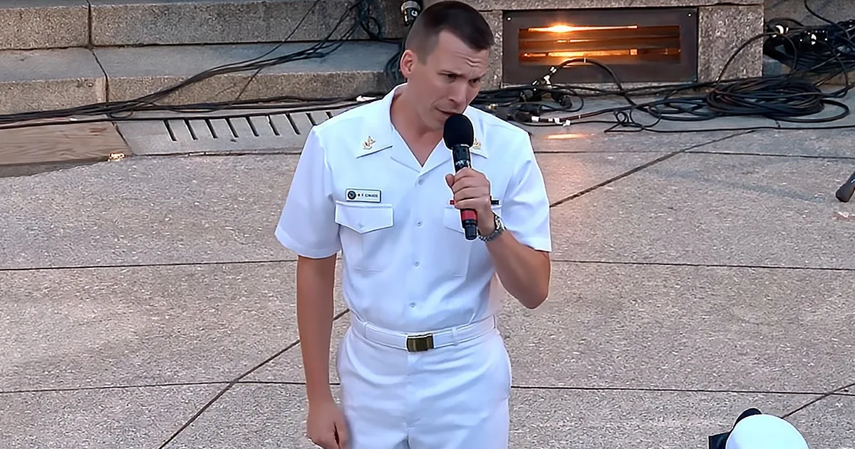 A man in a white naval uniform speaks into a microphone on a concrete surface.