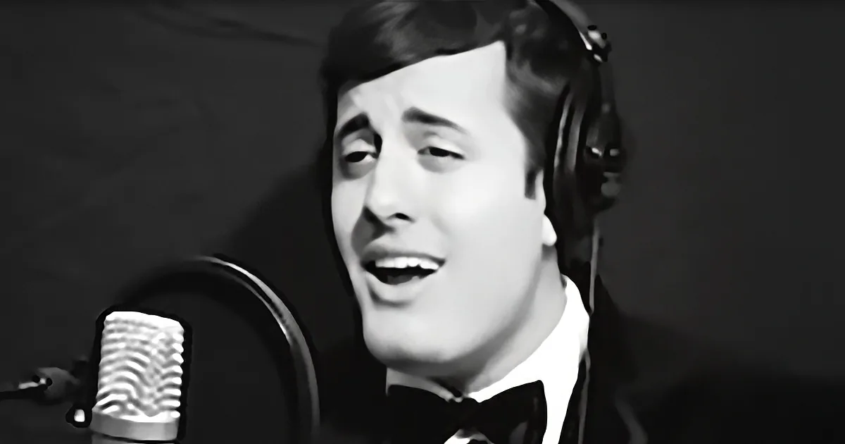 A man in a tuxedo sings into a microphone, wearing headphones, against a dark background.