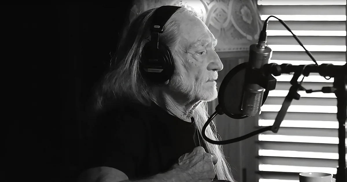 An older man with long hair and a beard is recording in a studio, wearing headphones and speaking into a microphone.