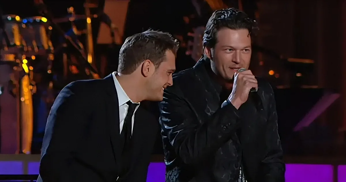 Two men, one singing into a microphone, the other smiling, on a stage with colorful lighting.