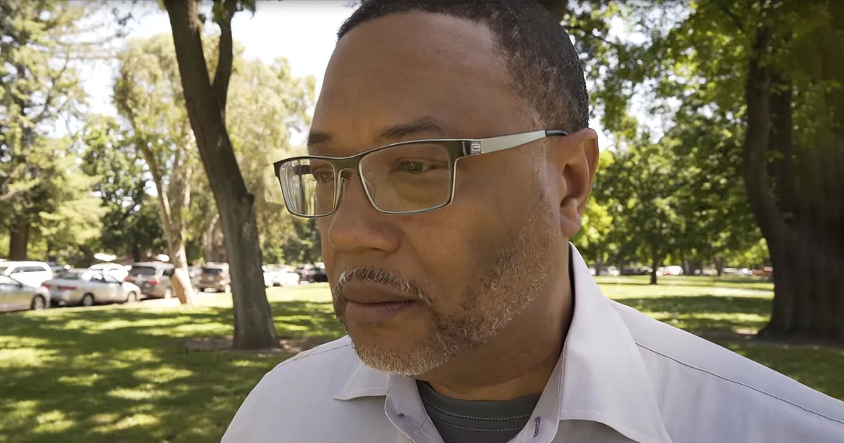 A man wearing glasses and a white lab coat stands in a park with trees and parked cars in the background.