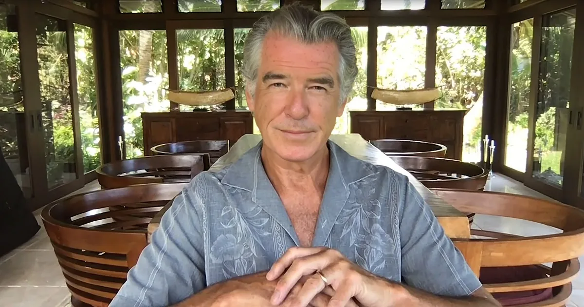 A man in a light blue floral shirt sits at a wooden table, hands clasped, in a sunlit room with large windows and...