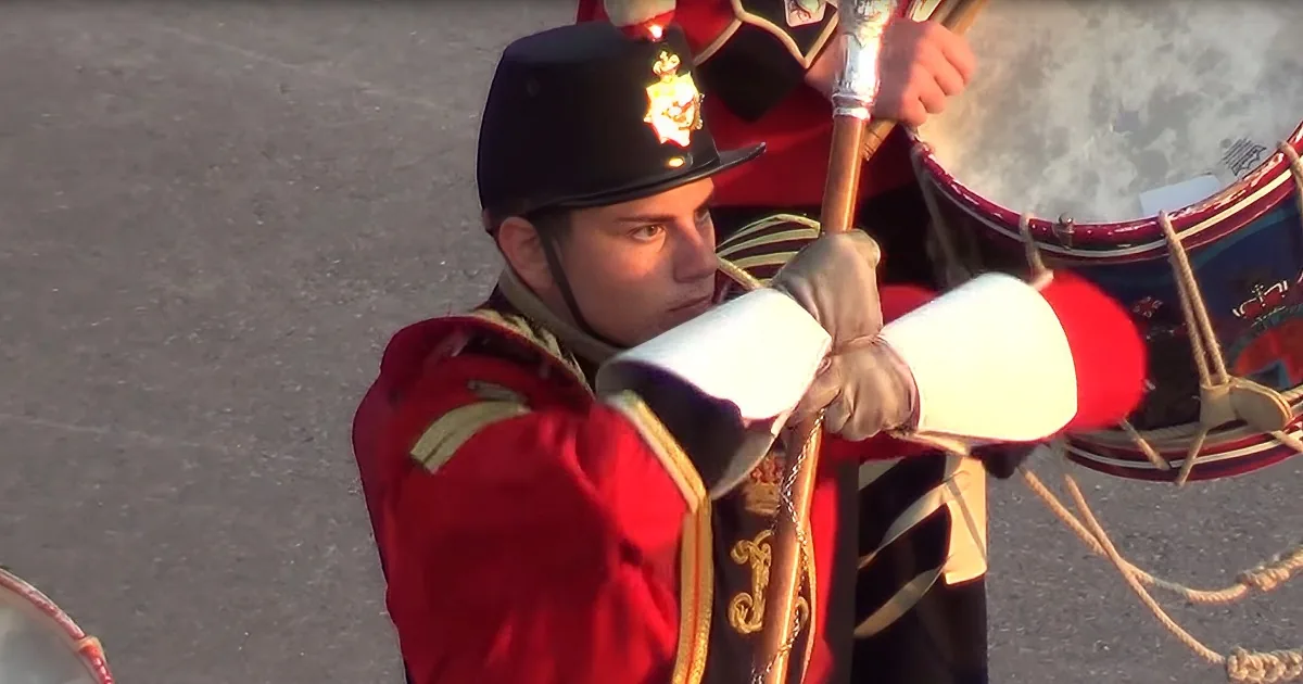A man in a red military uniform and black helmet plays a drum with white drumsticks.
