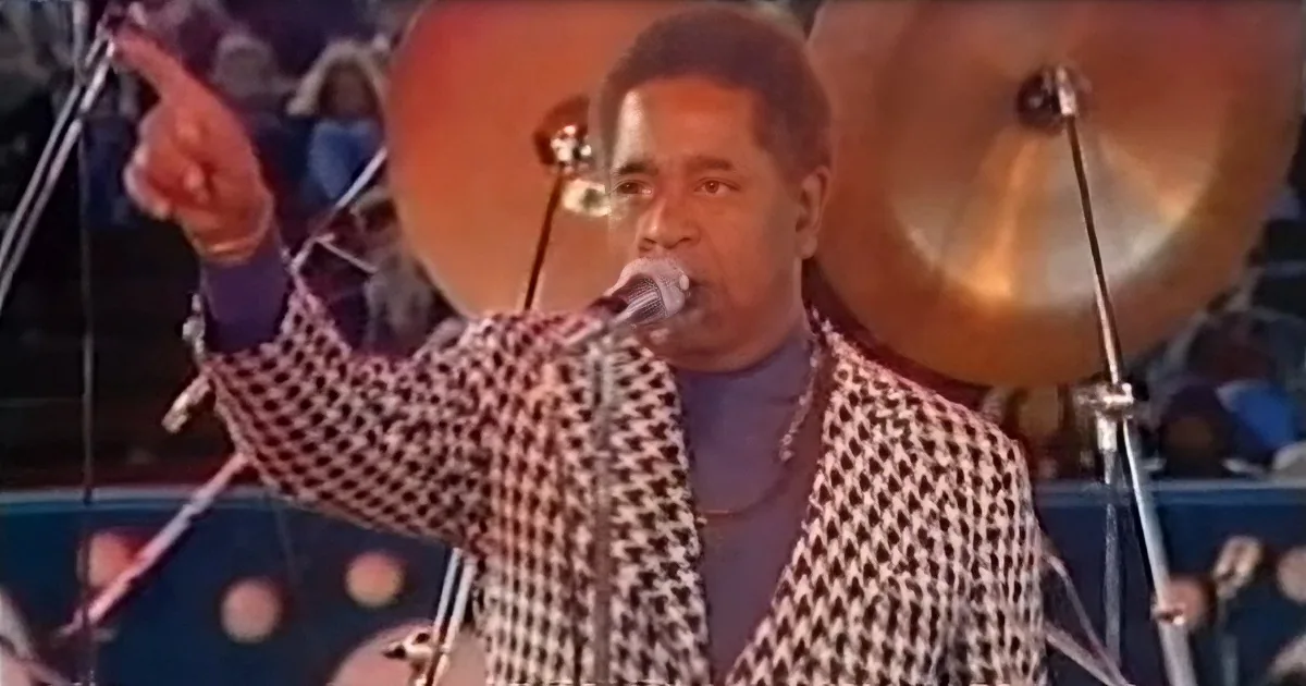 A man in a checkered blazer sings into a microphone, pointing to the left with a band in the background.