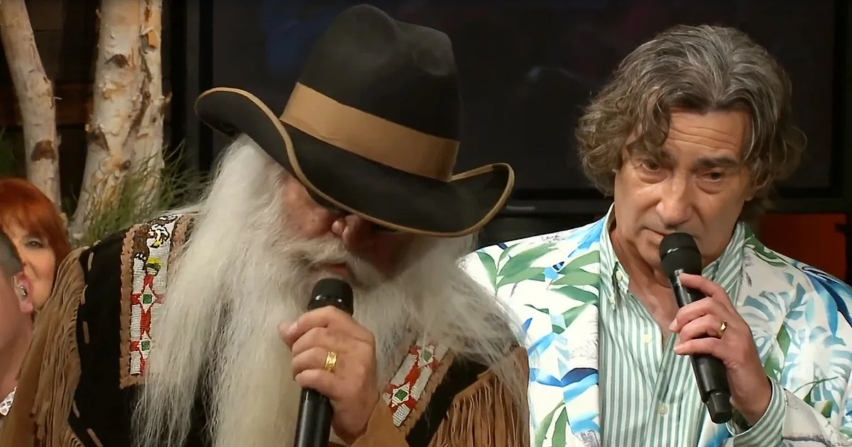 A man with a long white beard and hat holds a microphone, next to another man in a colorful shirt also holding a microphone.