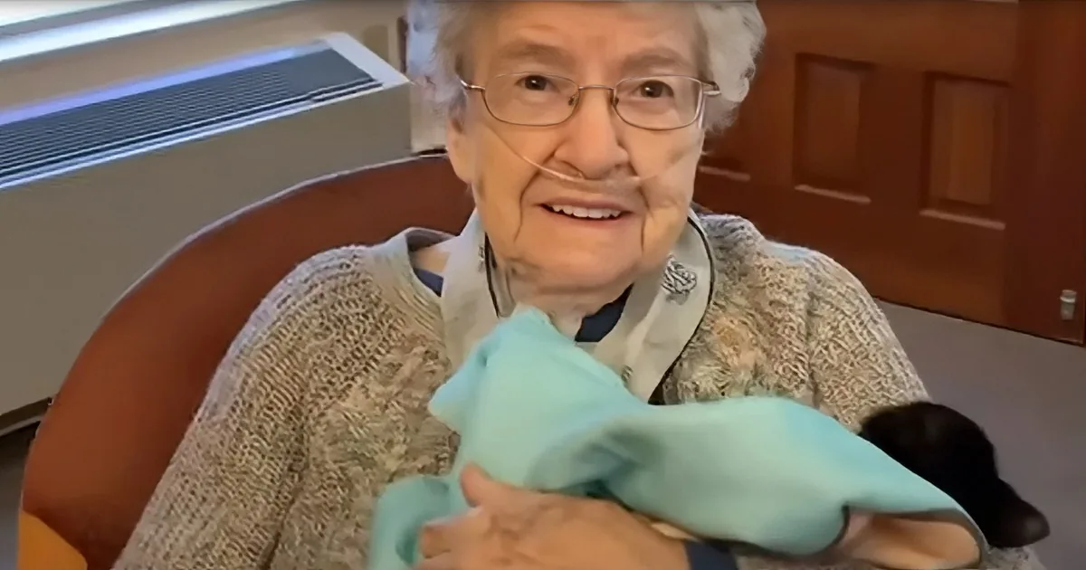 An elderly woman with glasses and a oxygen tube smiles while holding a light blue cloth.