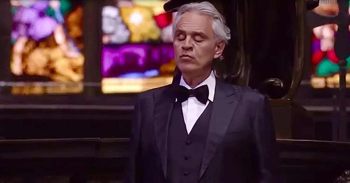 A man in a tuxedo stands in front of a stained glass window, eyes closed, wearing a bow tie.