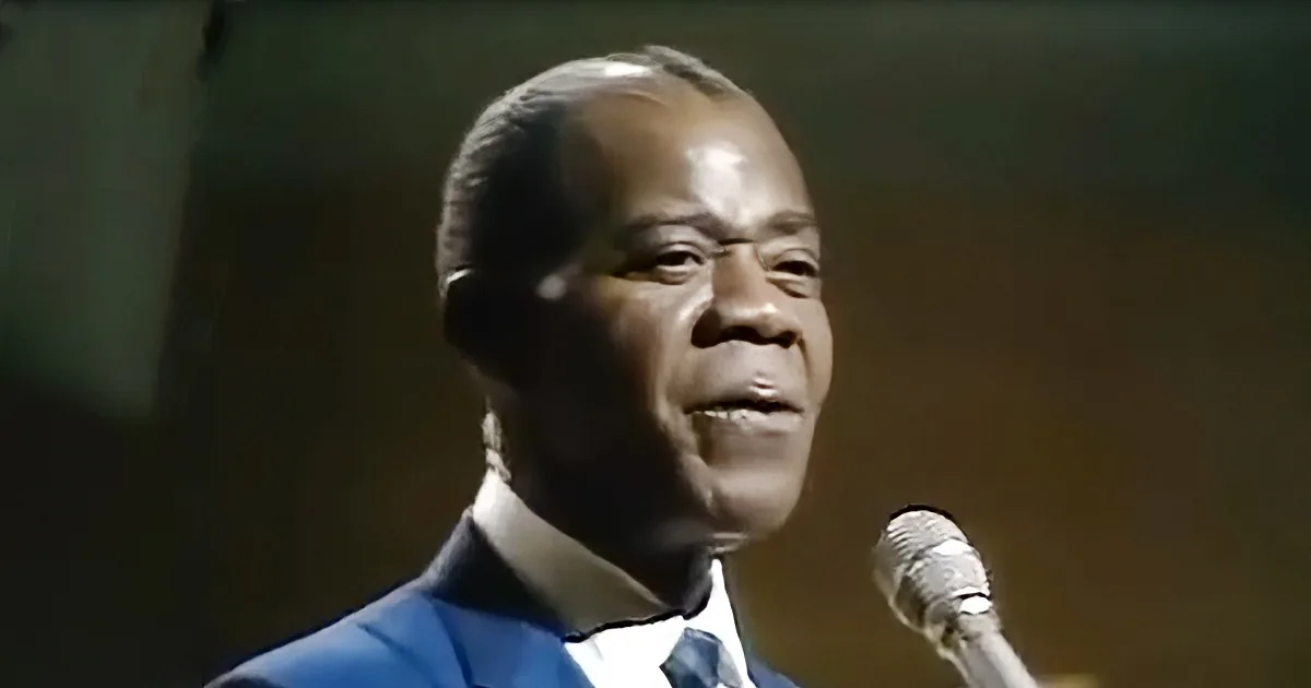 A man in a suit sings into a microphone, his face illuminated by stage lighting.