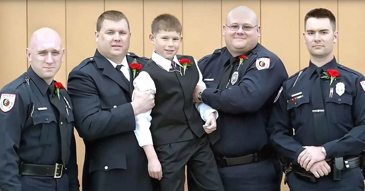 A young boy in a suit stands with four men in police uniforms, all wearing red rose boutonnieres.