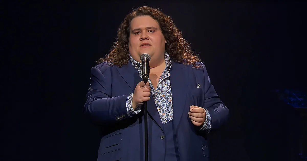 A man with curly hair in a blue suit and patterned shirt holds a microphone on stage.