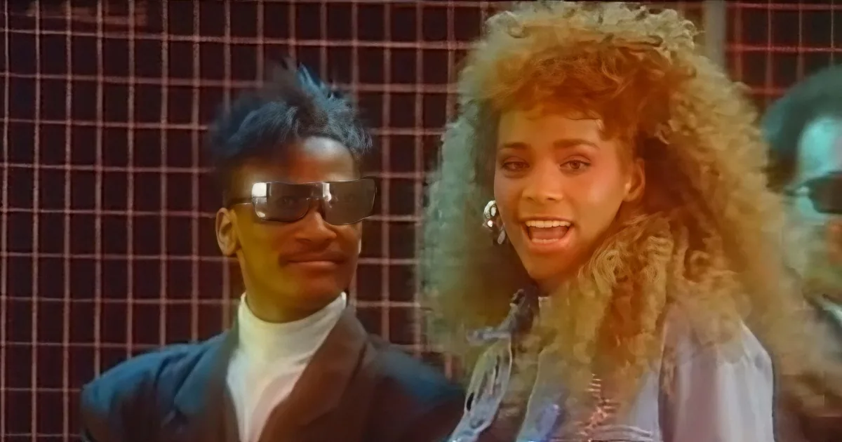 A man with blue hair and sunglasses stands next to a woman with voluminous blonde hair in a retro setting.