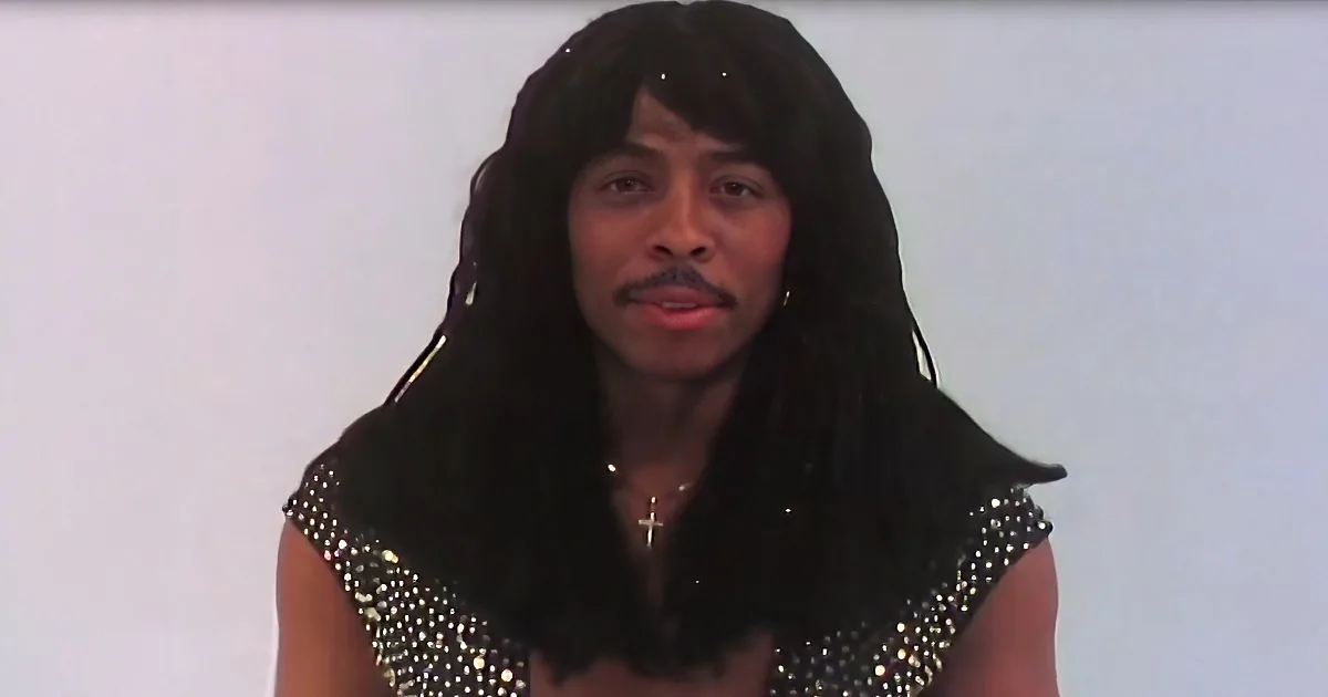 A person with long, dark hair and a studded top is wearing a cross necklace.