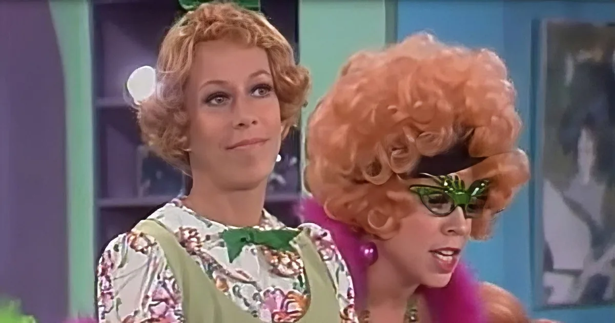 A woman with a large orange wig and green glasses stands next to another woman in a floral dress with a green bow.