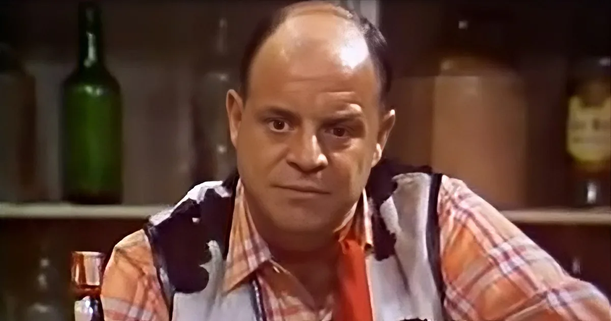 A man in a plaid shirt and suspenders sits at a bar, looking serious.
