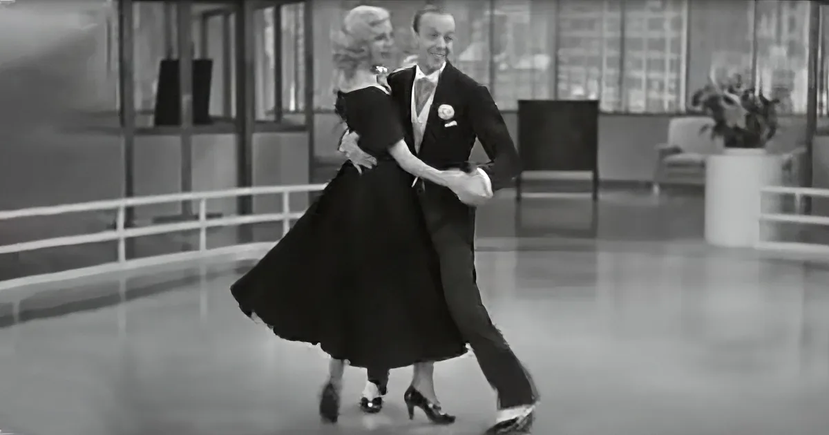 A couple in formal attire dances gracefully in a spacious room with a railing and potted plant.