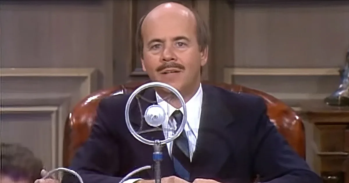 A man in a suit sits at a desk with a microphone, appearing to speak or listen intently.