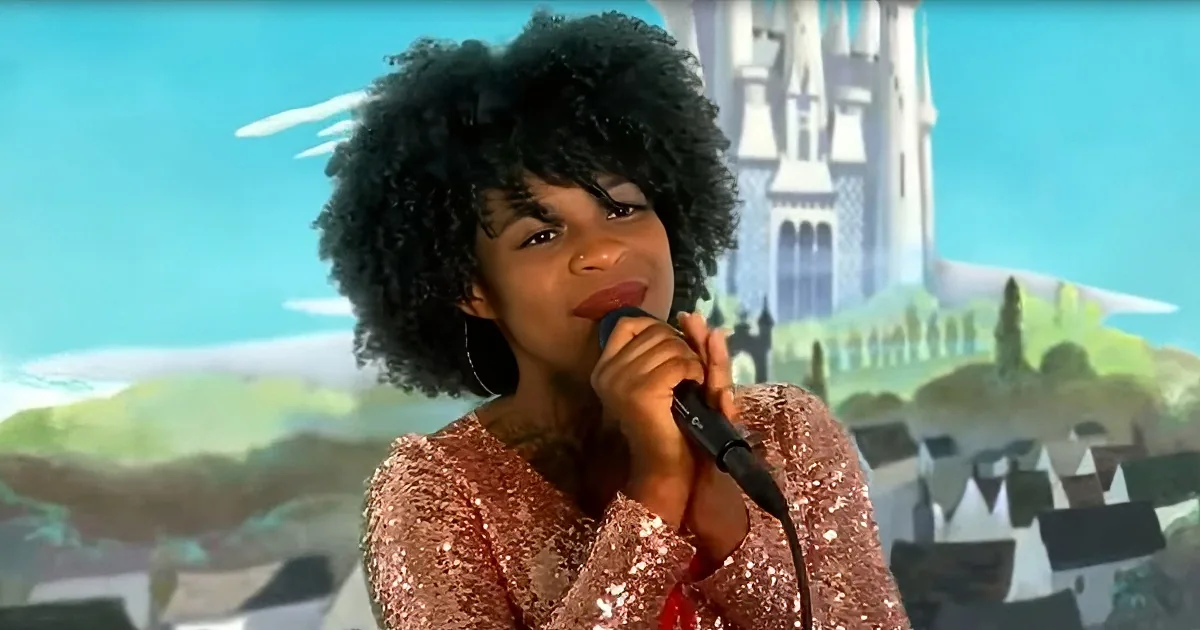 A person with curly hair in a sparkly pink outfit sings into a microphone against a castle backdrop.