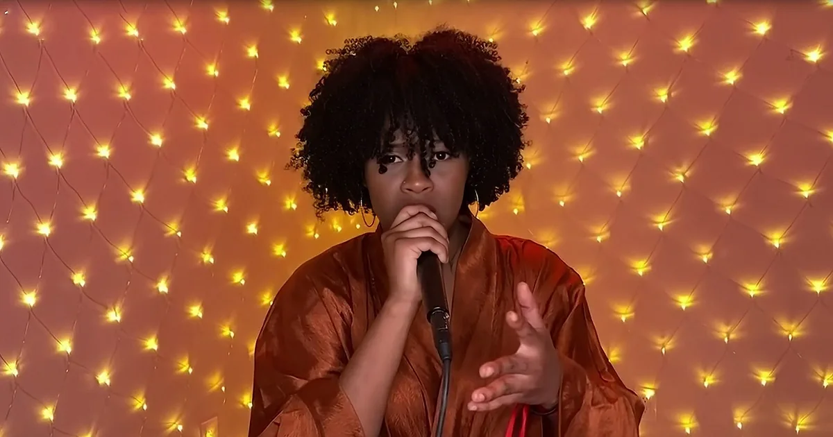 A person with curly hair in an orange outfit holds a microphone, gesturing with their other hand.