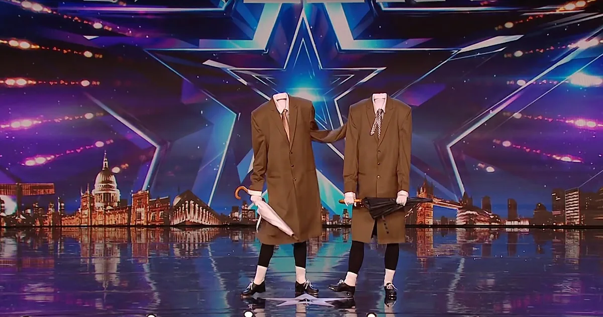 Two figures in brown suits with no heads, holding umbrellas, stand on a stage with a cityscape backdrop.